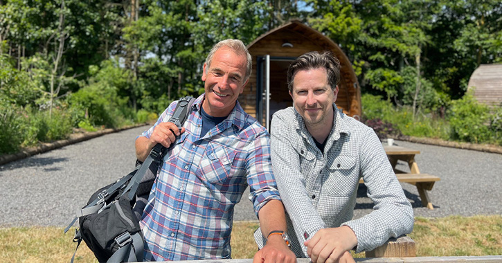 Robson Green and Lee Ingleby smiling at camera surrounded by green trees with glamping hut in the background.